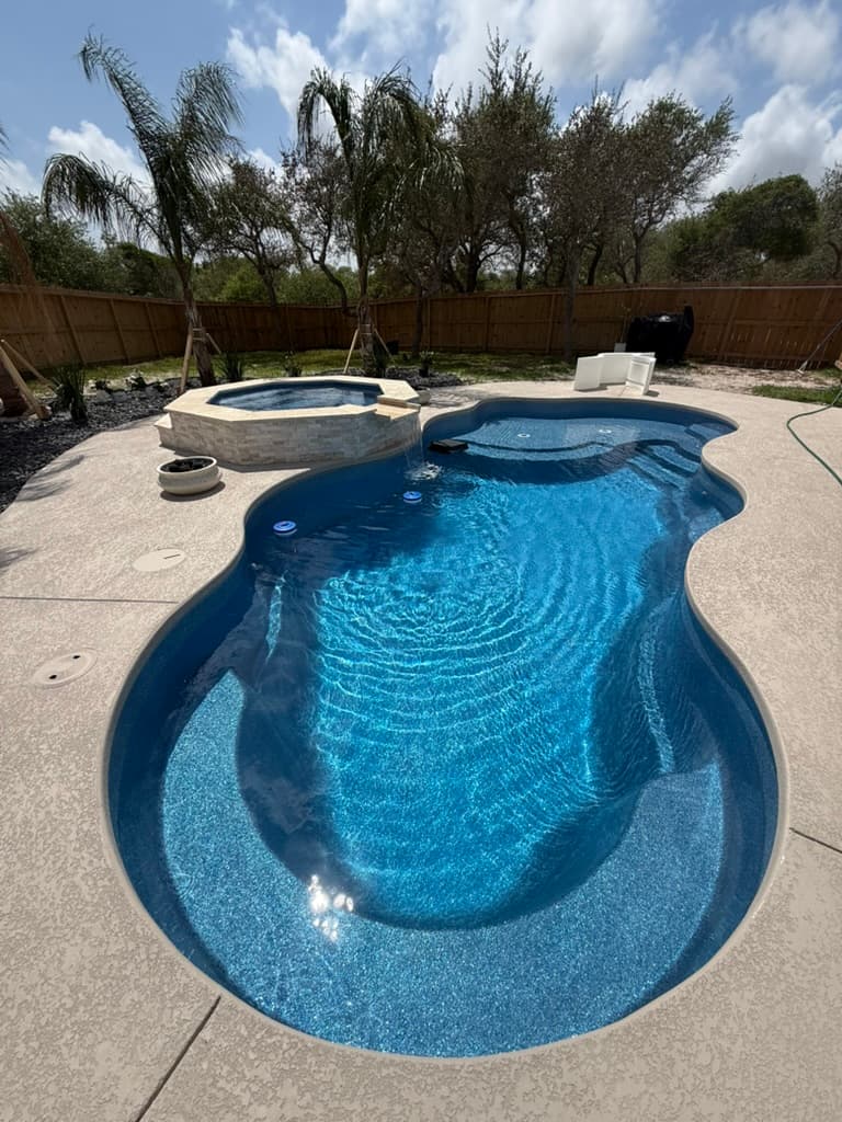 Light knockdown cool deck around a free-form fiberglass pool and raised spa, with a wood privacy fence and palm trees behind
