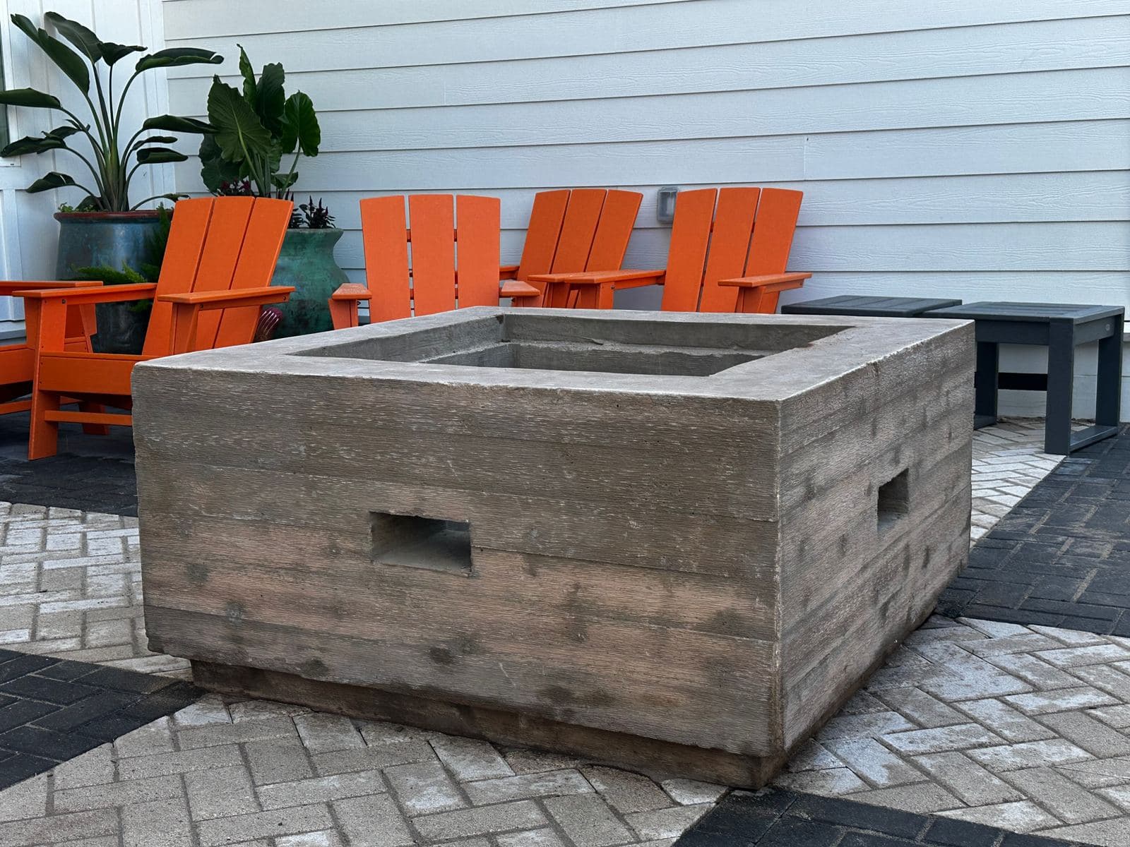 Square wood-grain concrete fire pit on a paver patio with orange chairs