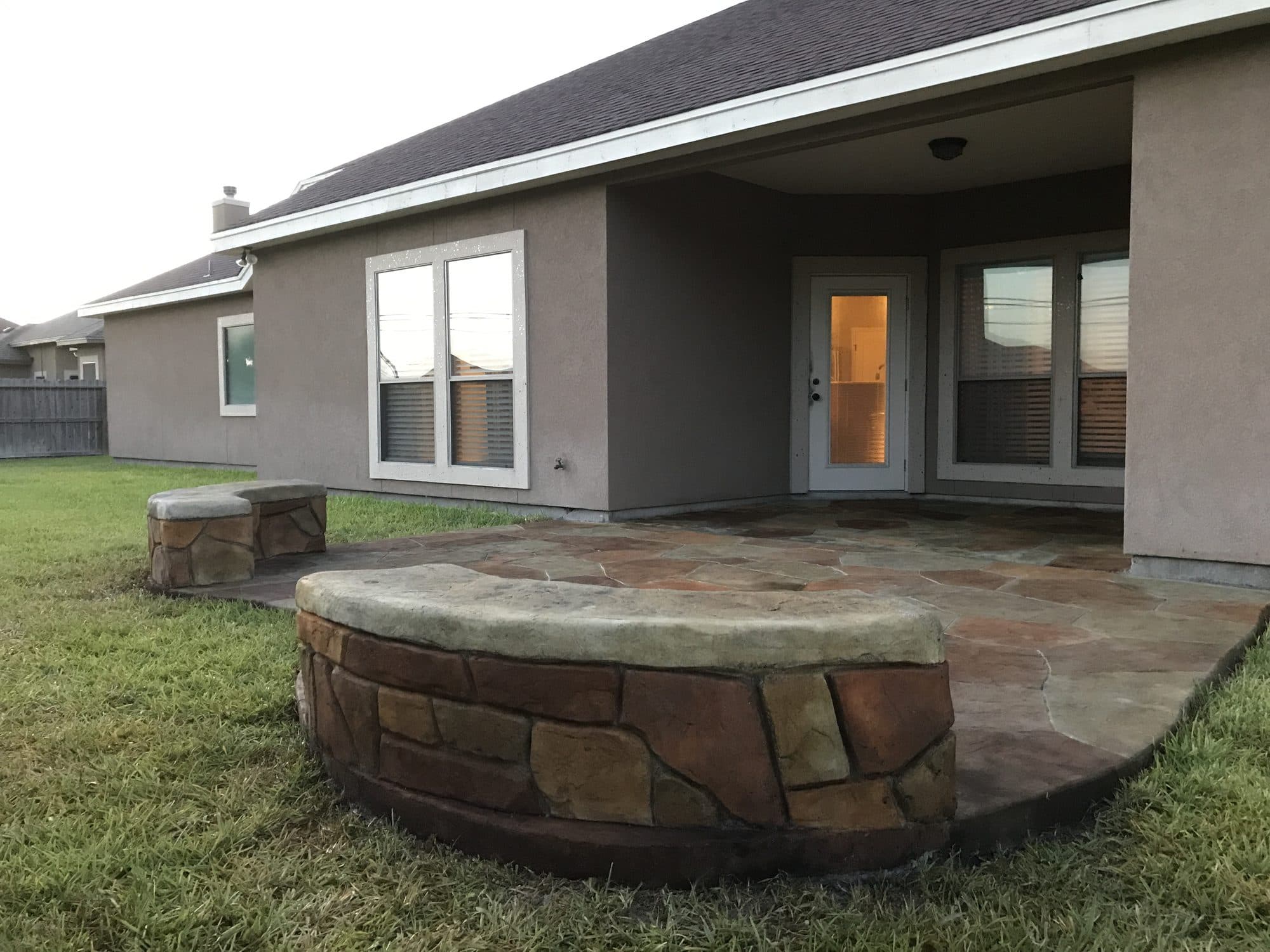 Round stacked-stone fire pit on a flagstone-look patio beside a stucco home