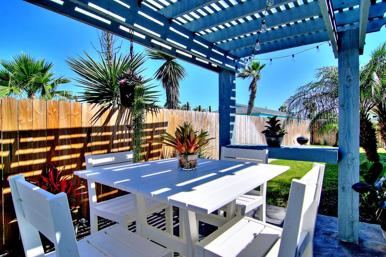 Blue pergola shading a white outdoor dining set beside tropical plants