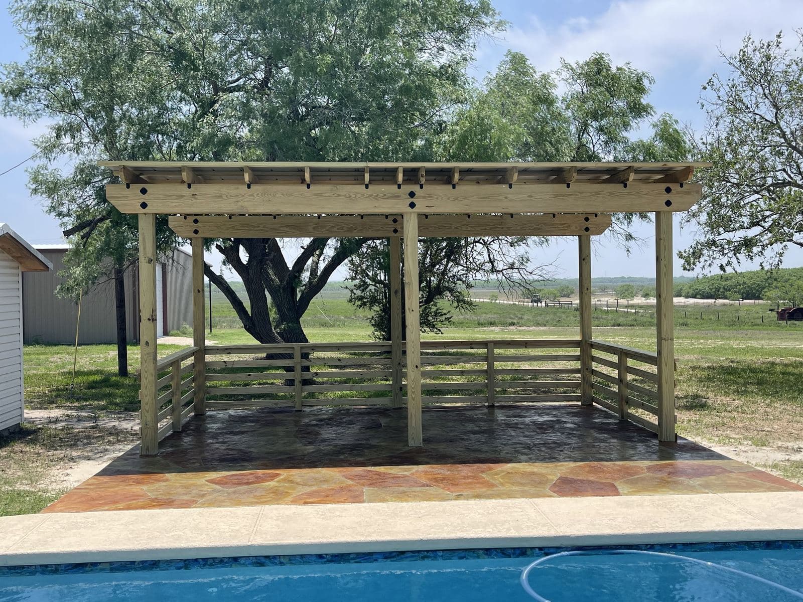 Pergola with wood railings over a stained stamped deck beside a pool